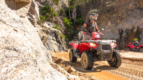 Helmeted rider on a red ATV splashing through sand on a sunny coastal canyon off-road trail beside rocky cliffs and palm trees