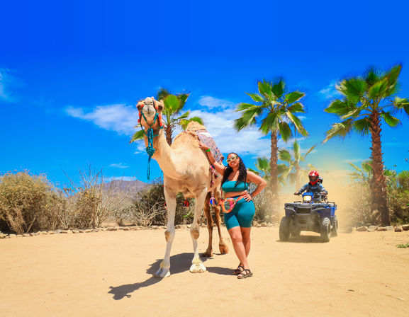 Woman in teal outfit posing with a saddled camel on a sunny desert oasis trail lined with palm trees while an ATV kicks up dust under a vivid blue sky — vacation camel ride and off‑road tour.