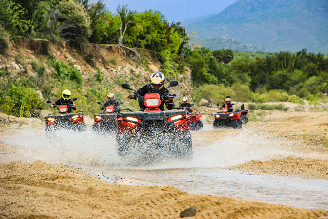 Group of riders on red ATVs splashing through a shallow stream on a sandy off-road desert trail with green scrub and mountains in the background.