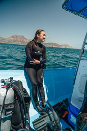 Smiling scuba diver in a black wetsuit with purple seams sitting on a bright blue dive boat, surrounded by tanks, fins and gear, turquoise open ocean and rocky coastal islands under a clear sky.