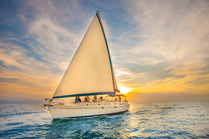 Sailboat cruising on calm open sea at golden sunset with people on deck, tall white sail and colorful sky reflected in gentle waves.