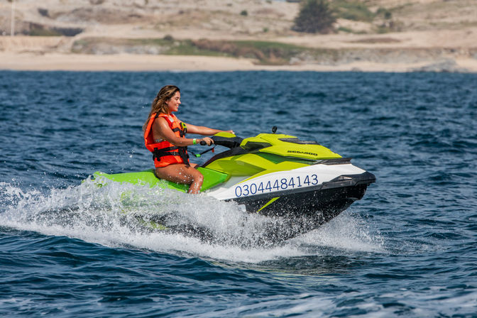 Rider in an orange life jacket on a lime-green jet ski skimming blue coastal waters near a sandy shoreline — energetic summer watersports scene.