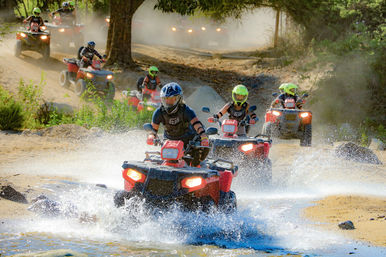 Group of helmeted riders on red ATVs splashing through a shallow stream on a dusty off-road trail, outdoor quad-bike adventure among trees and rocks.