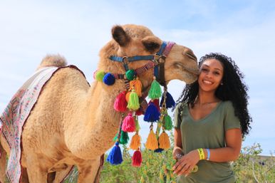 Smiling woman in a green shirt poses outdoors with a decorated camel wearing bright multicolored tassels and a patterned blanket under a sunny blue sky.