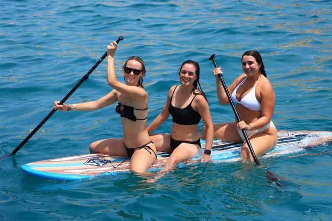 Three smiling women in swimsuits sharing a paddleboard and holding paddles on bright turquoise ocean water on a sunny day