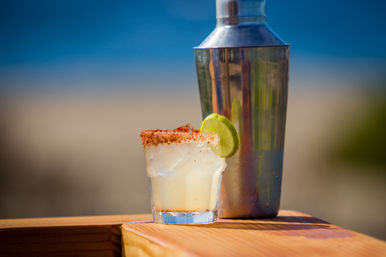 Bright lime cocktail with chili-salt rim and ice in a short glass, garnished with a lime wheel beside a metal shaker on a wooden railing with a blurred sandy beach and blue ocean background.