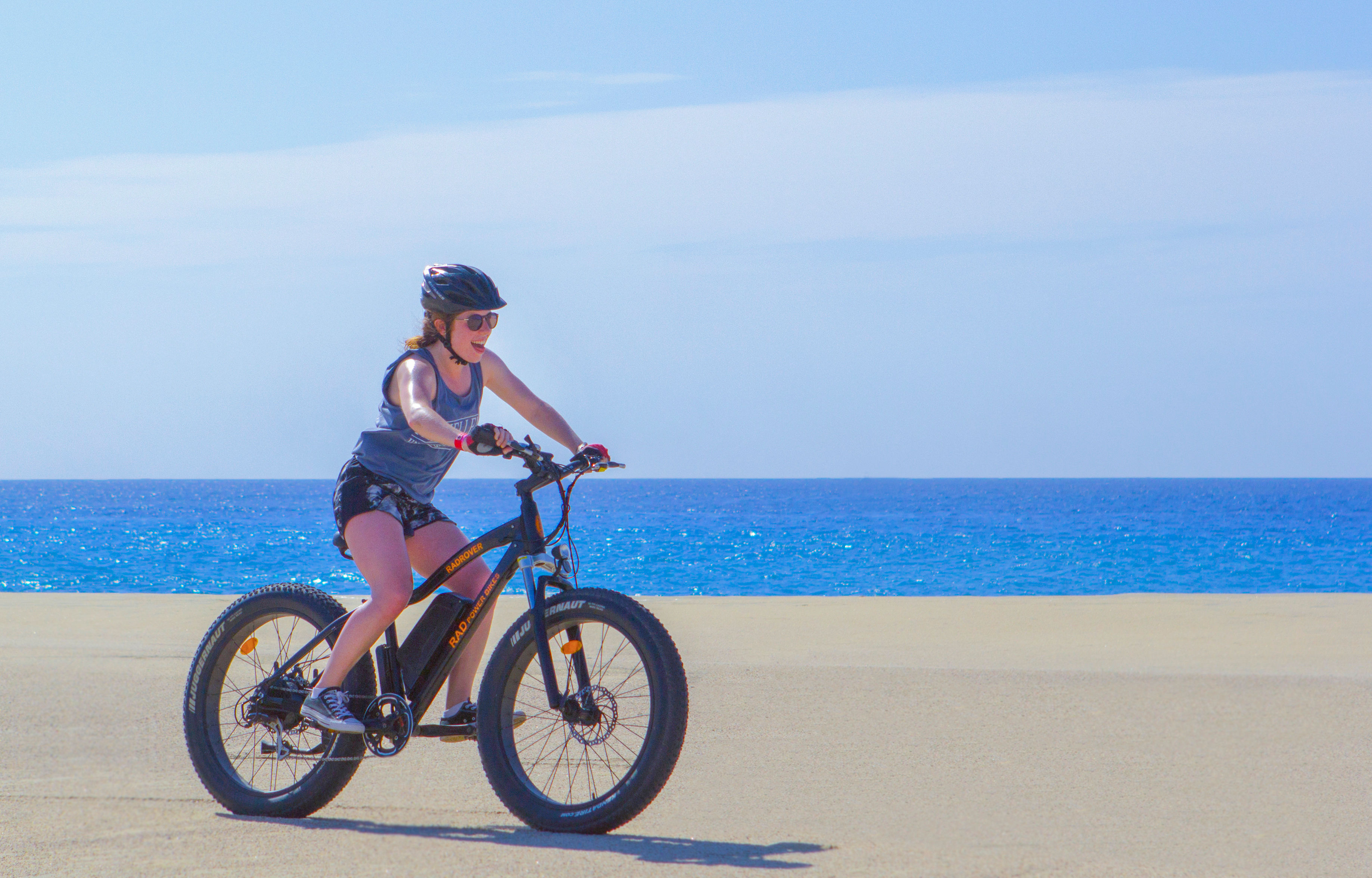 Cyclist in helmet and sunglasses riding a fat-tire e-bike on a sunny sandy beach with bright blue ocean and clear sky