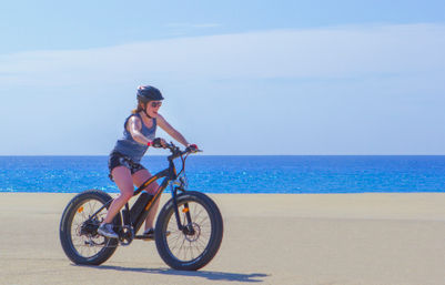 Cyclist in helmet and sunglasses riding a fat-tire e-bike on a sunny sandy beach with bright blue ocean and clear sky