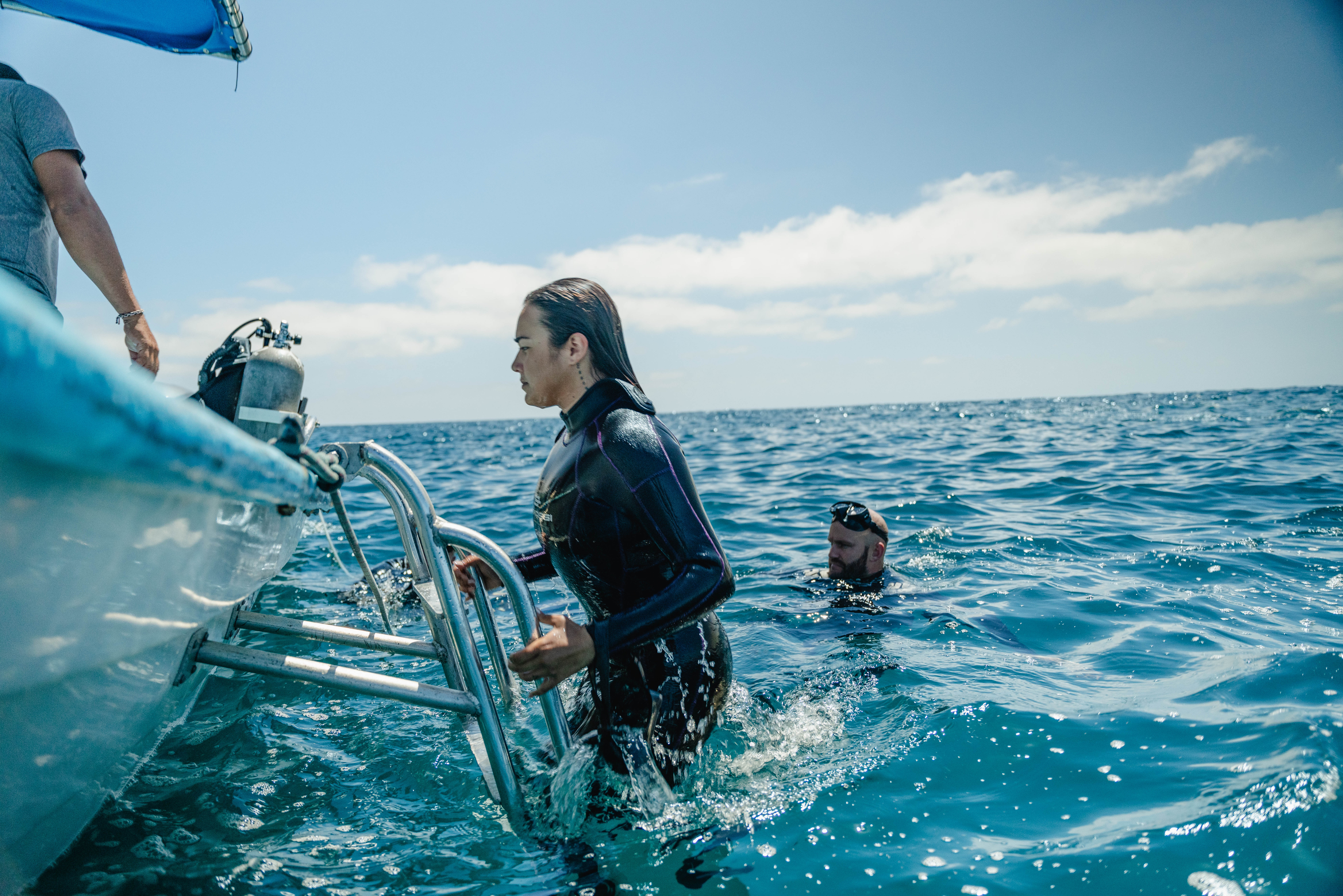Wetsuit-clad scuba diver climbing a boat ladder past a scuba tank while another diver floats in bright blue open ocean under a sunny sky