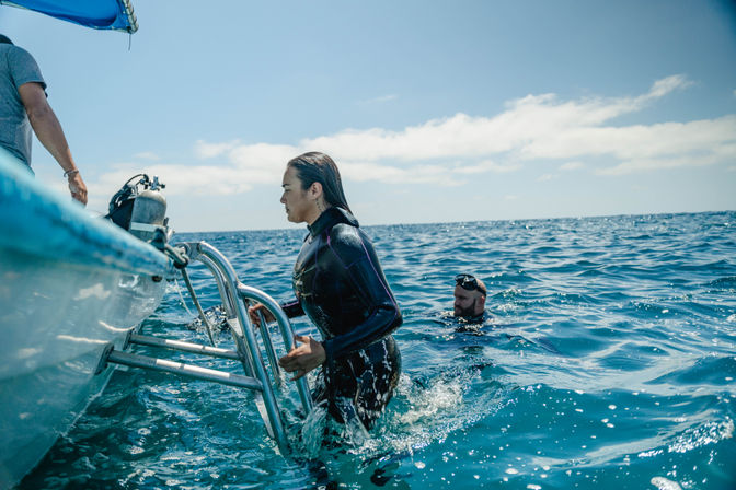 Wetsuit-clad scuba diver climbing a boat ladder past a scuba tank while another diver floats in bright blue open ocean under a sunny sky