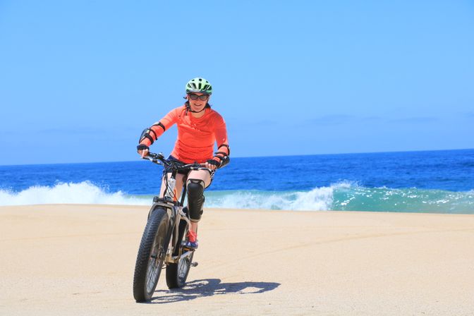 Smiling cyclist in an orange top and green helmet riding a fat-tire bike on a sunny sandy beach with blue ocean waves and a clear sky.