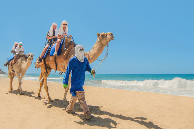 Tourists on camels led by a robed guide walking along a sunny sandy beach with turquoise waves and a clear blue sky.