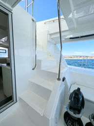 Sleek white yacht stairs with shiny stainless handrails leading to the upper deck, open cockpit overlooking blue coastal waters and shoreline under a clear sky, with a compact personal sea scooter docked nearby.