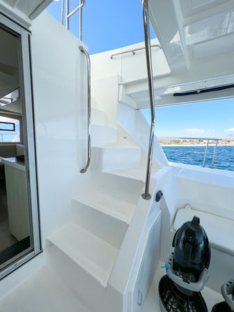 Sleek white yacht stairs with shiny stainless handrails leading to the upper deck, open cockpit overlooking blue coastal waters and shoreline under a clear sky, with a compact personal sea scooter docked nearby.