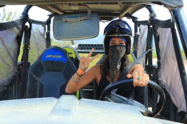 Off-road UTV driver wearing a helmet and bandana, flashing a peace sign while gripping the wheel on a dusty outdoor trail