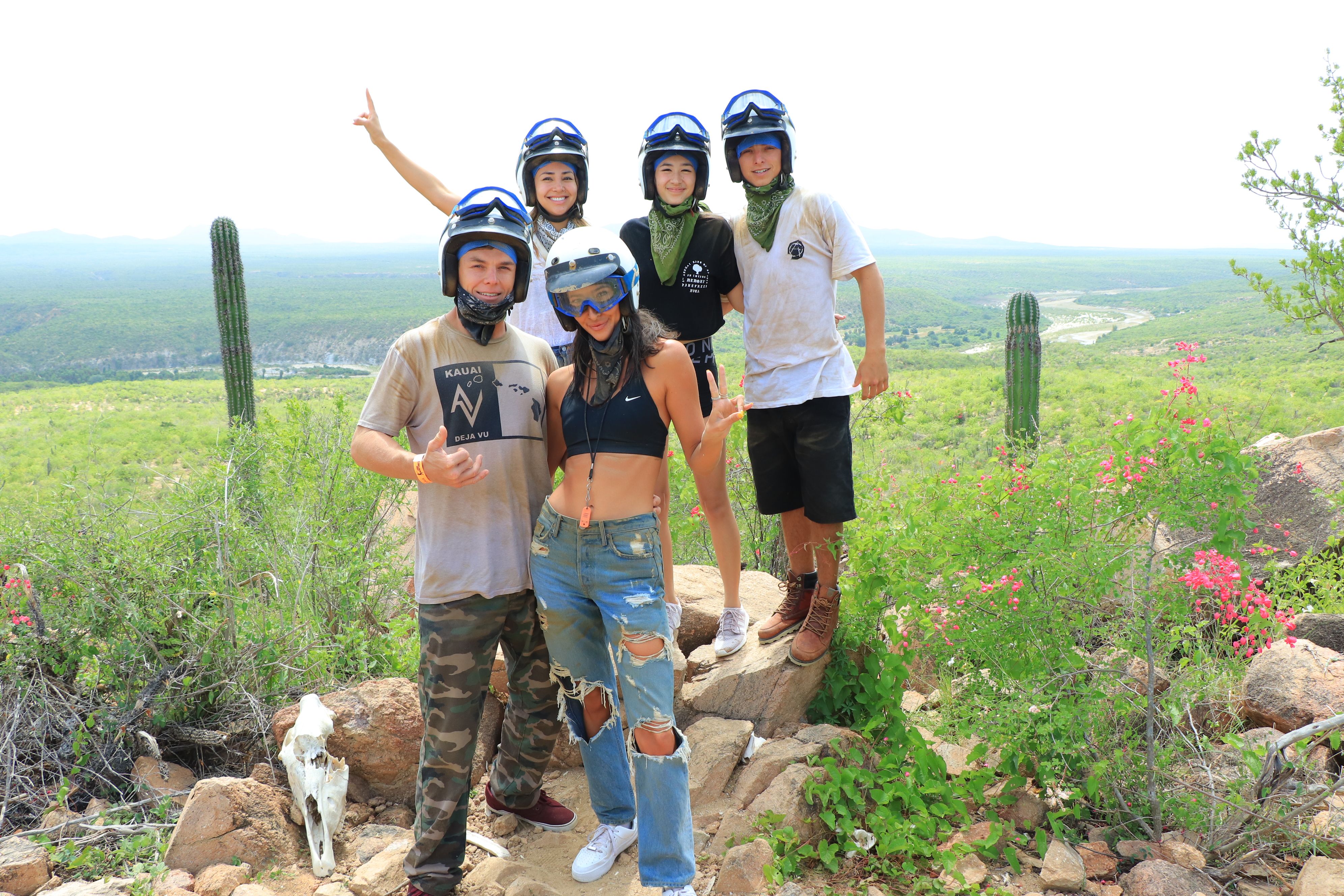 Group of five friends in helmets and bandanas posing on a rocky desert overlook with tall cacti and a green valley below after an off‑road adventure.