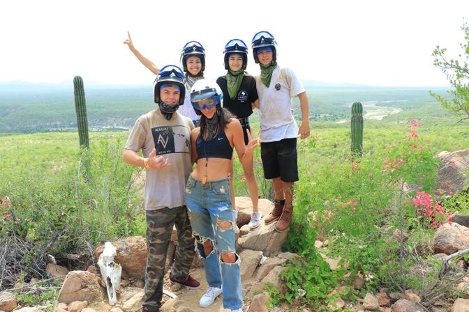 Group of five friends in helmets and bandanas posing on a rocky desert overlook with tall cacti and a green valley below after an off‑road adventure.