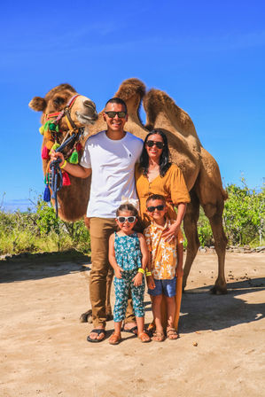 Smiling family of four in sunglasses posing with a two-humped camel wearing colorful tassels on a sunny outdoor desert tour under a bright blue sky