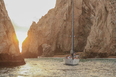 Sailboat with passengers sailing toward a dramatic coastal rock arch at sunset, golden light reflecting on calm ocean waters and towering rocky cliffs.