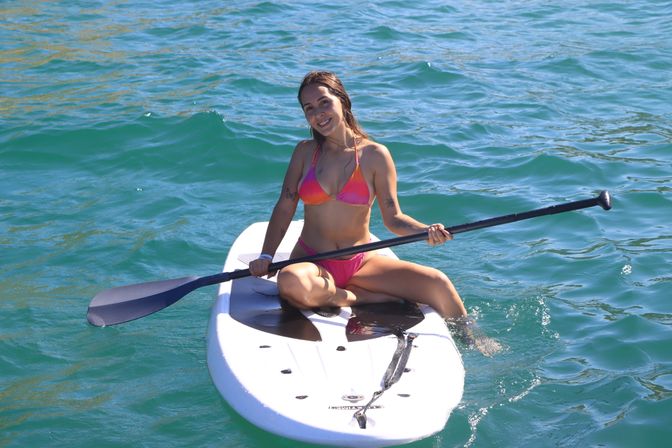 Smiling woman in a pink bikini sitting cross-legged on a white stand-up paddleboard, holding a paddle on turquoise water