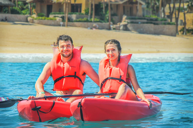 Two people in orange life jackets smiling in a red tandem kayak on clear blue water off a sandy tropical beach with palm trees