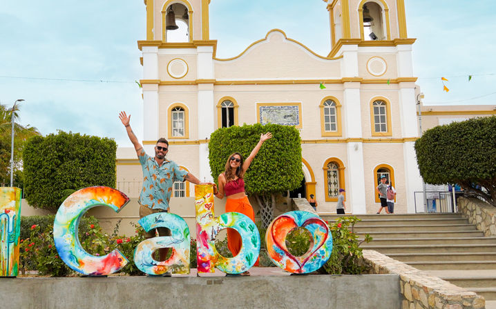 Two people waving beside colorful 'Cabo' letters and heart sculpture in front of a yellow-and-white colonial-style church in Cabo San Lucas, Mexico.