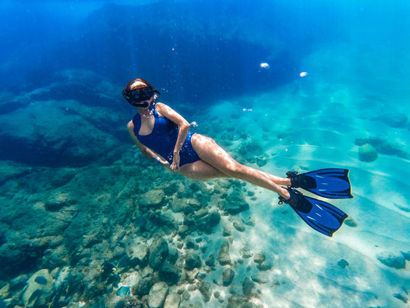 Snorkeler in a blue swimsuit and matching fins gliding above a rocky, turquoise seabed in clear tropical ocean water