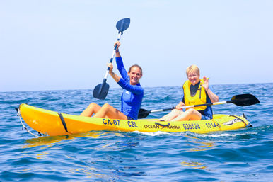 Two smiling paddlers in a yellow tandem ocean kayak on a sunny day—one raising a paddle and the other waving—gliding on calm blue sea while wearing life vests.