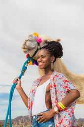 Smiling woman with braided updo holding a bright blue lead rope beside a camel decorated with colorful pom-poms, set against a cloudy desert sky and distant hills.