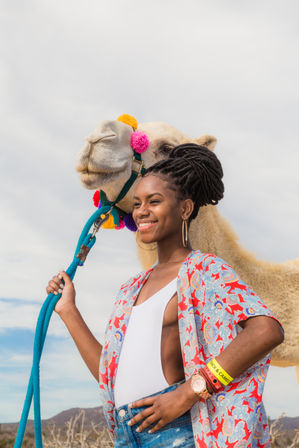 Smiling woman with braided updo holding a bright blue lead rope beside a camel decorated with colorful pom-poms, set against a cloudy desert sky and distant hills.