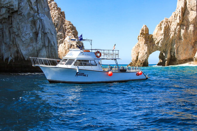 White scuba dive boat with orange buoys cruising turquoise ocean beside towering limestone sea arch and sunlit cliffs