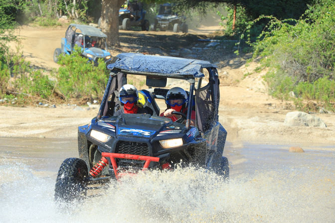Two helmeted riders in a blue off-road UTV splashing through water on a sunny dirt trail with scrubby vegetation