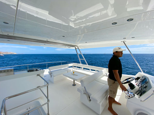 Sunlit luxury yacht deck with white lounge seating and table, crew member at the helm steering across deep-blue ocean toward a distant coastline under clear skies.
