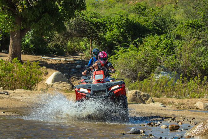 Two helmeted riders on a red ATV splashing through a shallow stream on a sunny off-road trail surrounded by green shrubs, rocks, and hillside — outdoor ATV adventure.