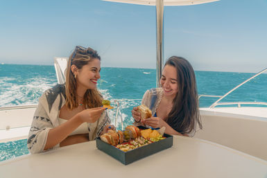 Two friends laughing and sharing sandwiches and snacks on a sunny yacht with turquoise ocean and rolling waves in the background.