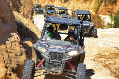 Convoy of off-road UTV side-by-side vehicles with helmeted drivers wearing green bandanas navigating a dusty, rocky canyon trail in bright sunlight