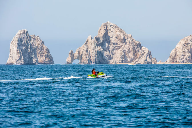 Rider on a bright green jet ski skimming deep blue ocean waters with a towering limestone sea arch and rugged coastal rock formations under a clear sky