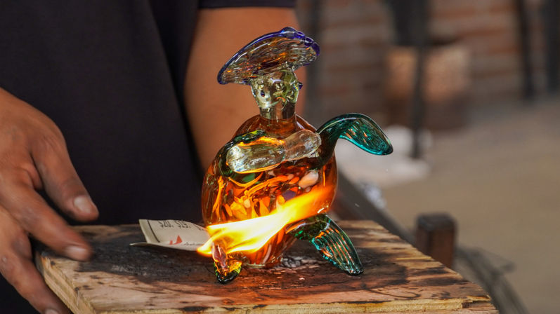 Vibrant orange-and-green blown-glass sea turtle glowing as a flame heats it on a charred wooden workbench, hands nearby in a glassblowing workshop.