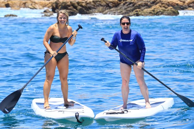 Two women smiling on stand-up paddleboards in clear blue ocean near a rocky shoreline, one in a black bikini and one in a blue rash guard, paddling on white boards.