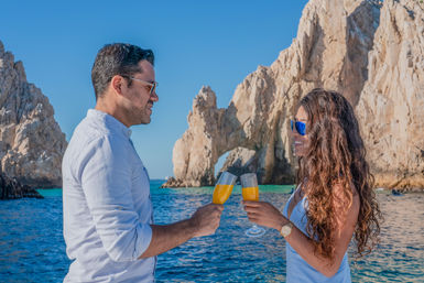 Sunlit couple toasting with orange cocktails on a boat beside a dramatic limestone sea arch and turquoise ocean — tropical vacation scene