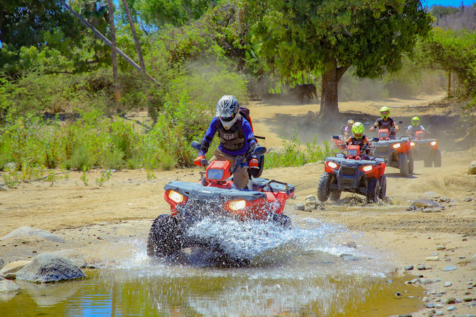 Off-road adventure: helmeted riders on red ATVs splashing through a shallow stream across a dusty riverbed lined with scrubby trees.