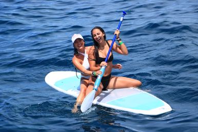 Two women in swimsuits laughing on a stand-up paddleboard (SUP) in deep blue ocean, one holding a paddle.