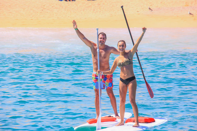 Smiling man and woman stand-up paddleboarding on turquoise ocean near a sandy beach, waving and holding paddles on a sunny day