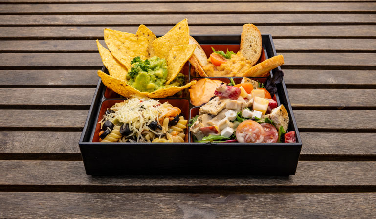 Vibrant bento-style black tray on a slatted wooden table with four compartments: tortilla chips and guacamole, creamy hummus with toasted bread, pasta salad with olives and grated cheese, and a mixed green salad with cherry tomatoes and diced chicken.
