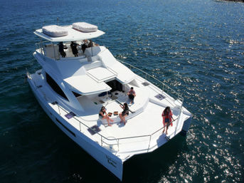 Aerial view of a white luxury catamaran on sparkling blue coastal water, people relaxing and sunbathing on the spacious foredeck under a sunny sky