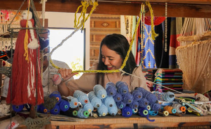Smiling woman at an artisan market stall handling braided yellow yarn amid stacked blue yarn spools, colorful woven textiles and hanging macramé decorations.