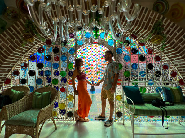 Silhouetted couple holding hands beneath a vibrant arched mosaic of colorful circular glass discs in a sunlit tiled indoor courtyard with wicker chairs and cushioned bench.