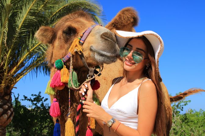 Smiling woman in a white sunhat and sunglasses posing with a colorfully tasseled camel by palm trees under a bright blue sky — sunny desert oasis vacation photo