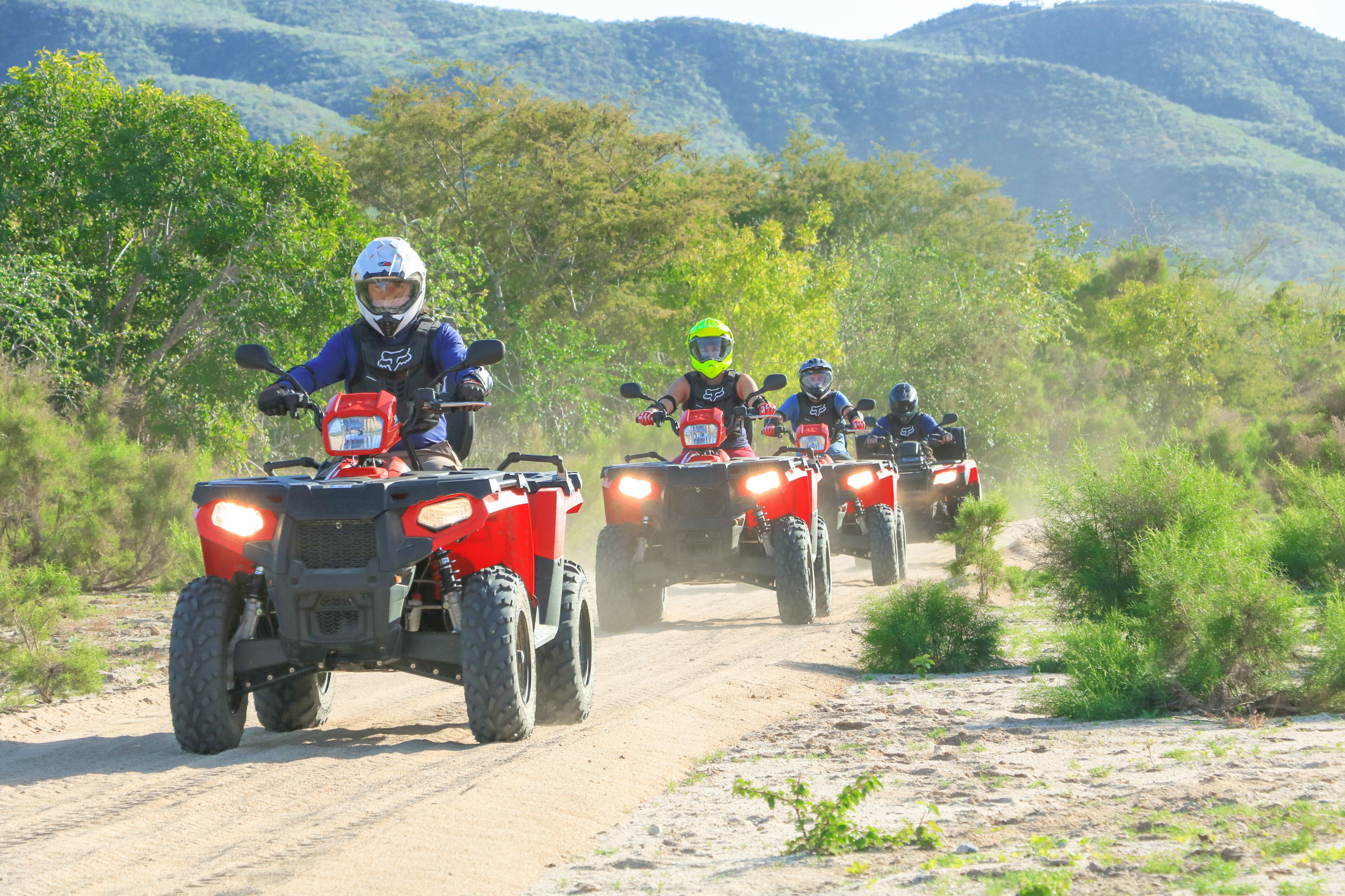 Group of four riders on red ATVs riding a dusty dirt trail through green scrub and mountain foothills — fun off‑road adventure in a scenic landscape