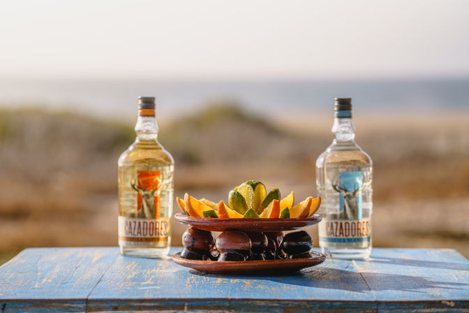Two clear tequila bottles flanking a clay dish of lime and orange wedges on a weathered blue wooden table with a blurred sandy beach and ocean in the background, seaside still life.